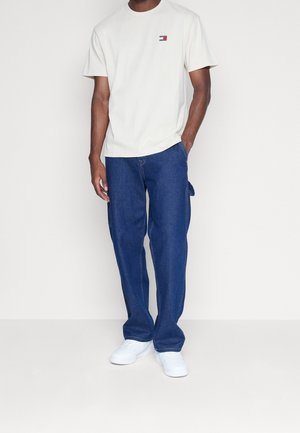 Lichtbeige korte mouw t-shirt met een klein logo op de borst, gecombineerd met losse blauwe spijkerbroek en witte sneakers.