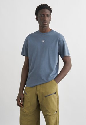 Man draagt een blauw The North Face T-shirt en mosterdkleurige cargobroek met ritszakken, poseert met één hand in de zak tegen een effen achtergrond.