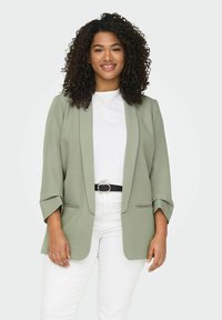 Světle zelený blazer se strukturovaným designem, otevřeným předním dílem, dvěma bočními kapsami a rukávy 3/4; nošený přes bílou košili a bílé kalhoty.