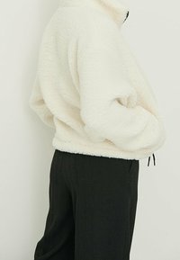 Pullover bianco in pile con colletto alto. Tessuto morbido e strutturato con vestibilità comoda, dotato di orlo regolabile e tasche laterali. Accenti neri visibili.