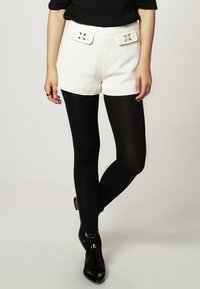 Vita högmidjade shorts med framfickor och dekorativa detaljer, parat med svarta täckande strumpbyxor och glänsande svarta ankelstövlar.