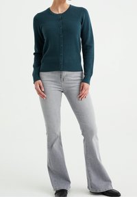 Teal knitted cardigan with a textured pattern and button front, paired with light grey flared jeans and black shoes.
