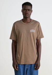 Camiseta de algodón marrón con cuello redondo y mangas cortas. En el frente presenta letras blancas con la palabra "DICKIES". Combinada con vaqueros azules.