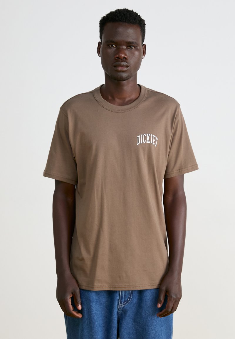 Camiseta de algodón marrón con cuello redondo y mangas cortas. En el frente presenta letras blancas con la palabra "DICKIES". Combinada con vaqueros azules.
