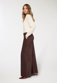 Pantaloni marroni a gamba larga con una texture morbida e tasche laterali, abbinati a un maglione color crema e tacchi rossi lucidi a punta.