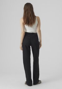 Jeans neri a vita alta con taglio dritto, dotati di due tasche posteriori e una consistenza denim robusta. Il modello indossa una canotta bianca aderente.