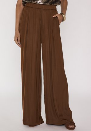 Vrouw die bruine hoge taille, wijd uitlopende broek draagt met hand in de zak, gecombineerd met bruine sandalen en gouden armbandjes om de pols.