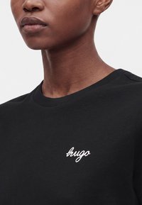 Gros plan d'une personne portant un t-shirt noir avec un logo blanc "hugo" sur la poitrine, montrant son cou et le bas du visage.
