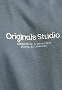 Grå bomuldsstof med glat tekstur, der har hvidt trykt tekst: "Originals Studio" og "Leveret til dig af Jack & Jones."