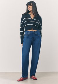 Melns izcirsts kardigans ar baltām svītrām, kombinēts ar zilām plašām džinsu biksēm un sarkanām spīdīgām plakanām kurpēm, uz neitrāla fona.