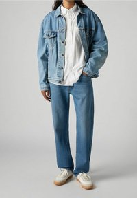 Chaqueta de mezclilla azul clara con cierre de botones, camisa blanca debajo, combinada con jeans azul claros y zapatillas blancas con suela beige.
