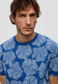 Niebieska koszulka z krótkim rękawem z białym floralnym wzorem i teksturalnymi detalami. Okrągły dekolt, luźny krój i miękki materiał.