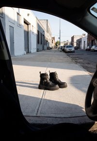 Een paar zwarte gespenlaarzen die op een zonovergoten stedelijke stoep staan, gezien vanuit een geparkeerde auto met gebouwen en auto’s langs de straat.