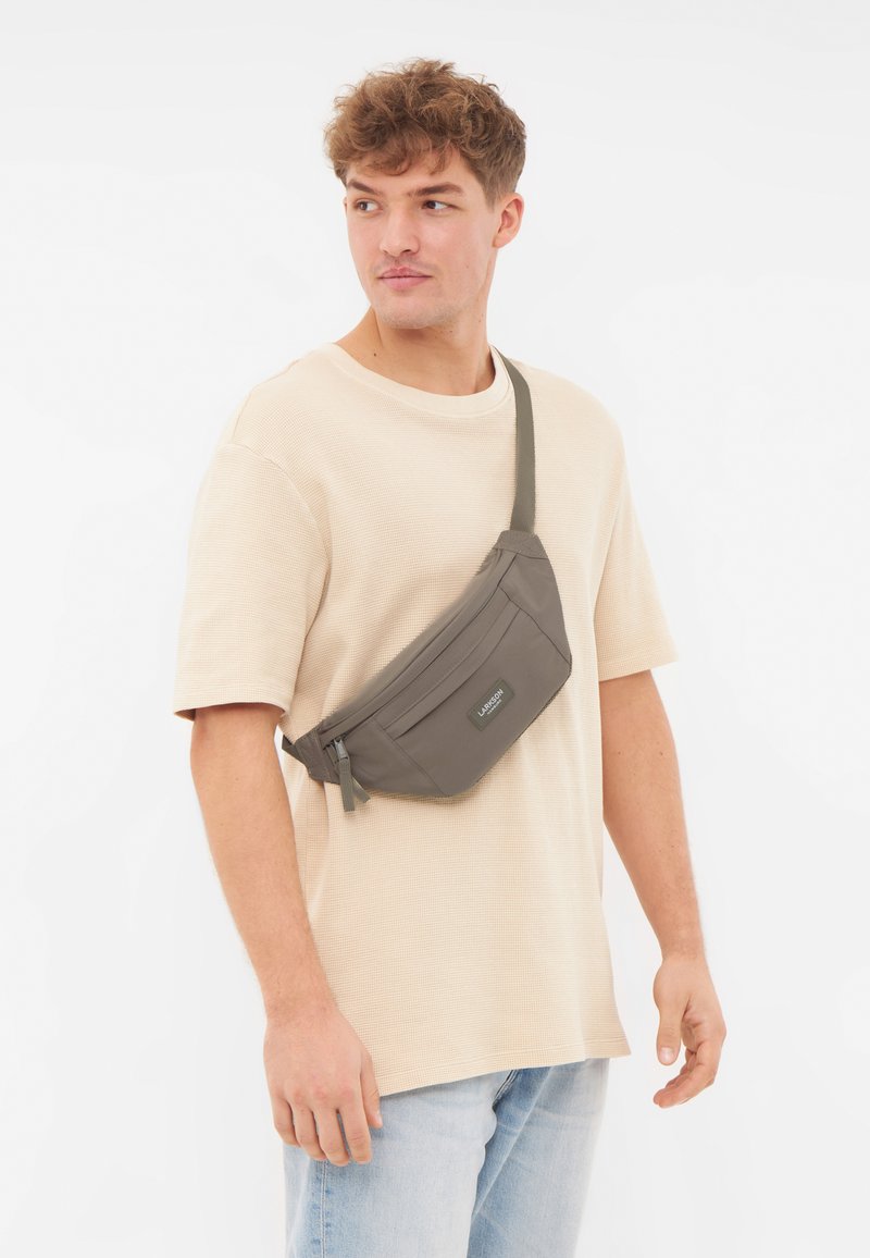 Borsa a tracolla grigia in nylon con una tasca con zip, indossata sopra una t-shirt beige con texture, dotata di una tracolla regolabile e un design minimalista.