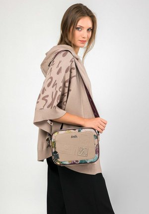 Junge Frau, die einen beigen, gemusterten Poncho und schwarze Hosen trägt, eine beige Umhängetasche mit bunten Kanten und geprägten Details trägt.