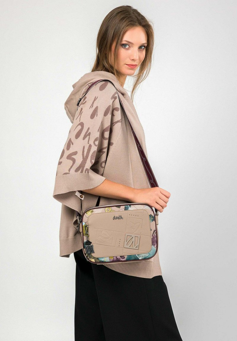 Junge Frau, die einen beigen, gemusterten Poncho und schwarze Hosen trägt, eine beige Umhängetasche mit bunten Kanten und geprägten Details trägt.