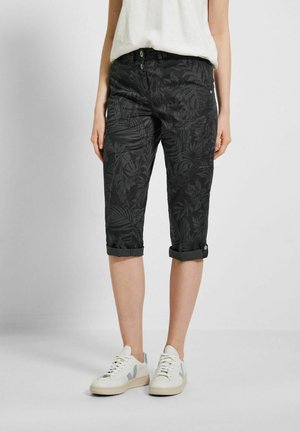 Kvinde iført opkørte sorte cropped bukser med mørke bladmønstre, hvid ærmeløs top og hvide sneakers med grå accenter.