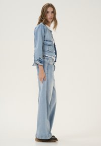Jachetă din denim și blugi cu picior larg în albastru deschis. Jacheta are buzunare și margini desfăcute; blugii au o croială relaxată și detalii de custură.