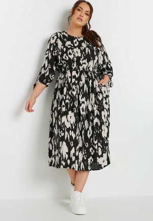 Vestido midi de estampado de leopardo en negro y crema, con mangas tres cuartos, cintura elástica, bolsillos laterales y una falda fluida, combinado con zapatillas blancas.