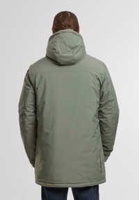 Giacca isolante verde con cappuccio, caratterizzata da una superficie liscia, un taglio dritto e tasche laterali con zip. Vista posteriore mostrata.