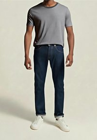 Camiseta gris de manga corta con cuello redondo, vaqueros azul oscuro, zapatillas blancas. Los vaqueros tienen un diseño clásico de cinco bolsillos y detalles de costura.