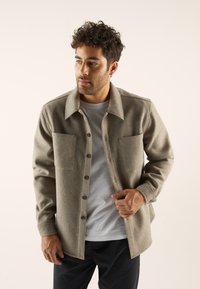 Veste chemise en mélange de laine grise avec un devant boutonné, deux poches poitrine et un col classique, portée avec un t-shirt blanc en dessous.