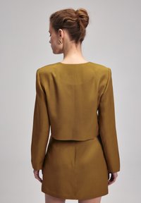 Kort, senapsgul jacka med långa ärmar och beskuren design, tillsammans med en matchande minikjol, visad bakifrån.