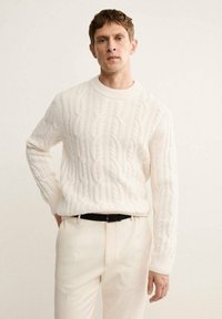 Kremowy sweter w splot warkoczowy z okrągłym dekoltem, z wzorzystą fakturą, w połączeniu z jasnymi spodniami i czarnym paskiem.