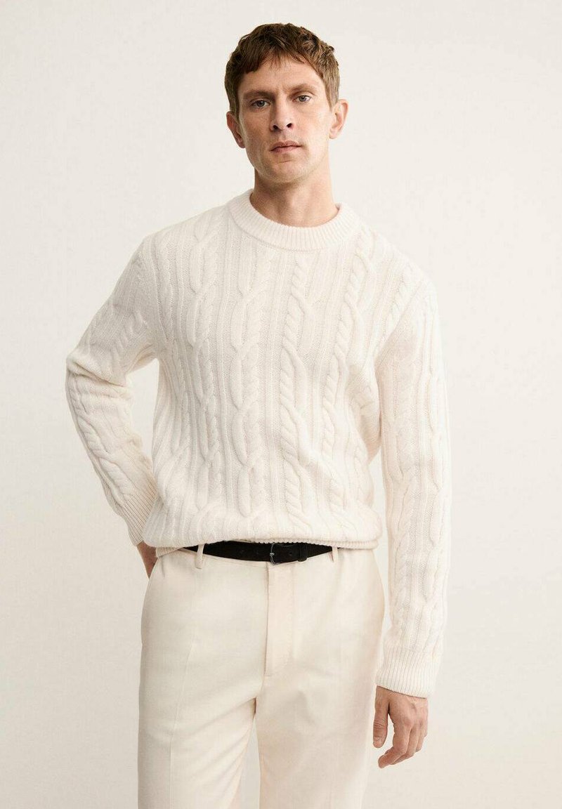 Cream cable-knit sweater with a round neckline, featuring textured patterns, paired with light trousers and a black belt.