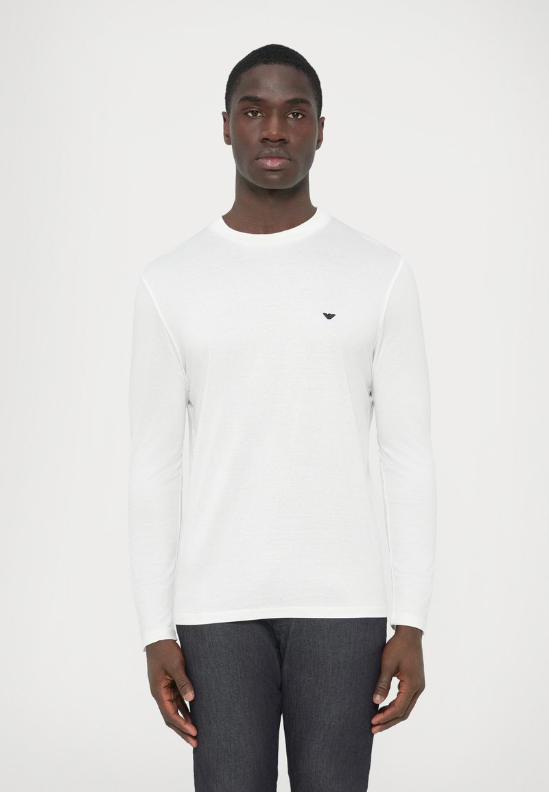 Άσπρο πουκάμισο με μακριά μανίκια και στρογγυλή �λαιμόκοψη, κατασκευασμένο από λείο ύφασμα. Διαθέτει διακριτικό μαύρο λογότυπο στο πάνω αριστερό μέρος του στήθους.