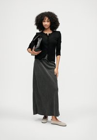 Črni cardigan, siva maksi krilo z vezalko in bež natikači. Modelka drži črno torbico in stoji na nevtralnem ozadju.