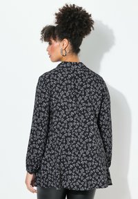 Schwarzes Shirt mit langen Ärmeln und lockerer Passform, verziert mit einem weißen Blattmuster. Der Stoff hat eine glatte Textur und einen subtilen Glanz.