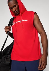 Red sleeveless hoodie with a front kangaroo pocket, featuring white "Champion" text across the chest and drawstrings on the hood.