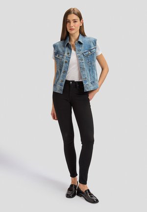 Fiatal nő áll, kék ujjatlan farmermellényt, fehér pólót, fekete szűk nadrágot és fekete mokaszint visel, egyszínű háttér előtt.