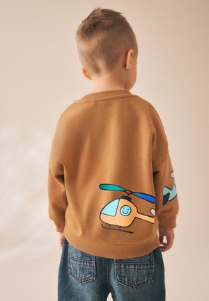 Barn set bagfra iført en brun sweatshirt med en smilende tegneseriehelikopter og blå jeans mod en beige baggrund.
