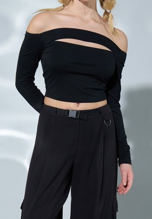 Černý zkrácený top s dlouhým rukávem, off-shoulder designem a výřezy. V kombinaci s černými kalhotami s vysokým pasem, které mají pásek a kovové detaily.