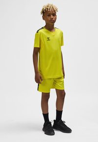 Żółta sportowa koszulka i krótkie spodenki wykonane z gładkiego materiału, z czarnymi akcentami na ramionach i logo. Noszone z czarnymi skarpetkami i trampkami.