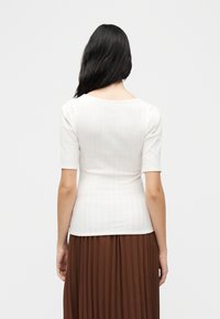 Γυναίκα με μακριά σκούρα μαλλιά που φοράει μια εφαρμοστή λευκή ριπ μπλούζα με κοντά μανίκια και καφέ πλισέ φούστα, εμφανιζόμενη από πίσω σε λευκό φόντο.