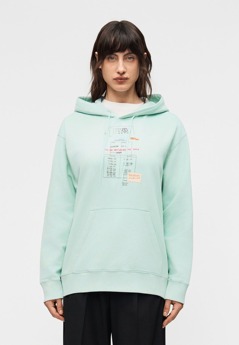 Γυναίκα με σκουρόχρωμα μαλλιά που  φοράει γκριζοπράσινο φούτερ με κουκούλα, με τυπωμένο ημερολόγιο και χειρόγραφες σημειώσεις, στέκεται μπροστά σε μια λευκή απλή φόντο.