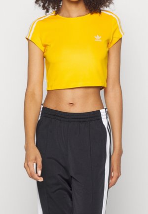 Kobieta ubrana w żółtą krótką koszulkę Adidas z białymi paskami na rękawach oraz czarne spodnie dresowe z białymi paskami po bokach.