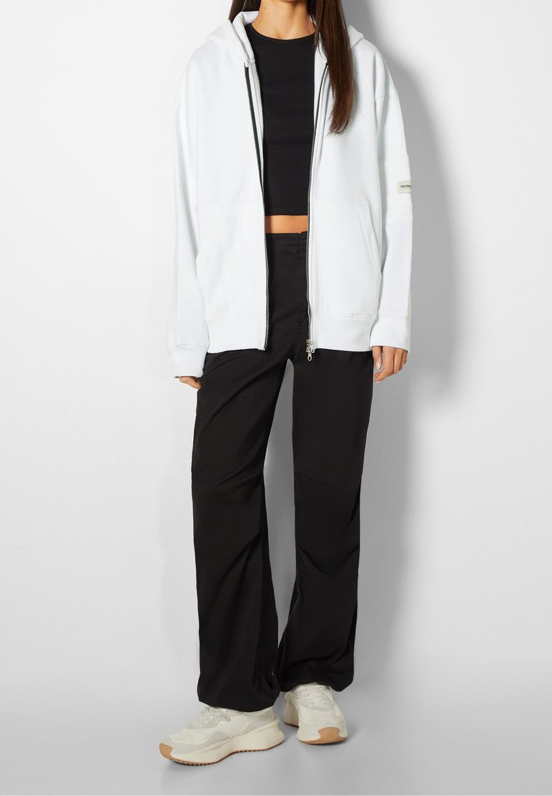 Femme portant un sweat à capuche blanc zippé, un crop top noir, un pantalon large noir et des baskets blanches, debout devant un fond blanc uni.