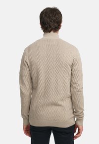 Suéter de punto beige con un patrón de espiga texturizado, cuello alto y dobladillo y puños acanalados, combinado con pantalones oscuros.