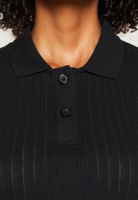 Närbild av en person som bär en svart ribbad piké med två svarta knappar fastknäppta vid kragen.