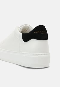 Baskets en cuir blanc avec un accent en daim noir au talon et un logo doré. Semelle blanche épaisse avec des détails texturés et des perforations sur la tige.