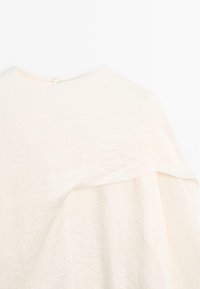 Crèmekleurig textiel kledingstuk met een gestructureerd oppervlak, met een gelaagd ontwerp en een knoopsluiting bij de achterkant van de halslijn.