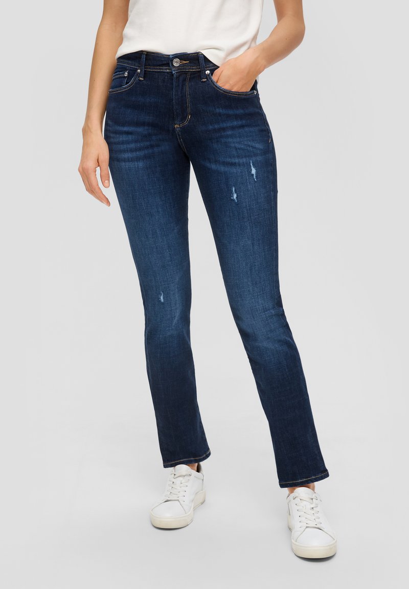 Jean en denim bleu foncé au design ajusté, légèrement déchiré à l'avant, et un style classique à cinq poches, porté avec des baskets blanches.