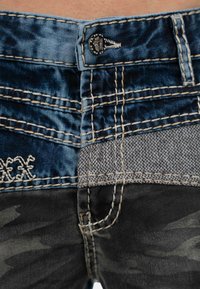 Shorts en denim avec un design mixte de motifs de camouflage bleu foncé, gris et noir, ornés de coutures contrastantes et d'un détail de bouton décoratif.