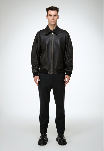 Hombre de pie mirando hacia adelante con una chaqueta de cuero negra, pantalones negros y zapatos gruesos negros sobre un fondo claro y simple.