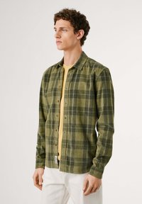 Grønn rutet skjorte med brune knapper, lange ermer, krage-design, og en gul undertrøye, laget av teksturert stoff.