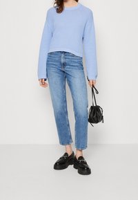 Žena oblečená v světle modrém pleteném svetru, vysokých modrých džínách, černých robustních mokasínech a držící malou černou tašku na šňůrku.